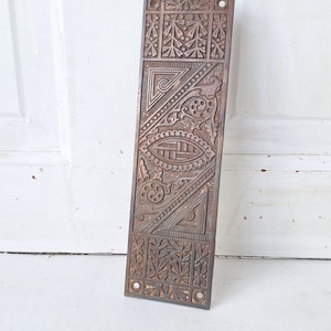 May include: A rectangular, antique bronze door plate with intricate, symmetrical patterns. The design includes floral and geometric shapes, with a central oval element. The plate has a weathered appearance and small holes for mounting.