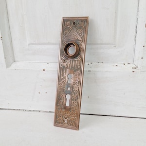 May include: An antique bronze door plate with intricate embossed patterns. The rectangular plate features a circular opening for the doorknob and keyholes. The design includes geometric shapes and floral motifs, showcasing a vintage aesthetic.