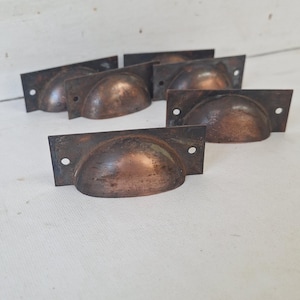 May include: A collection of antique bronze-colored cup pulls. Each pull has a rectangular backplate with two screw holes and a rounded, cup-shaped handle. The pulls are arranged on a white surface.