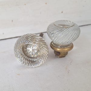 May include: Two clear glass door knobs with a spiral design. The knobs have a brass base and are set against a white background. The glass has a textured, ribbed pattern, and the knobs are likely vintage or antique.