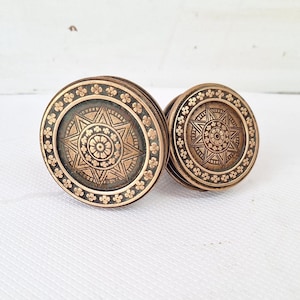May include: Two antique bronze cabinet knobs. Each knob features a central starburst design surrounded by a floral border. The knobs have a circular shape and a warm, aged patina, suggesting they are vintage or vintage-inspired.