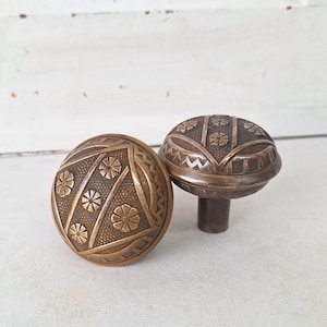 May include: Two antique bronze door knobs with floral and geometric patterns. The knobs are round with a stem on one, and are set against a white background.