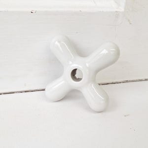 May include: A white ceramic faucet handle with a cross-shaped design. The handle has four rounded arms extending from a central hole. The glossy finish reflects light, highlighting its smooth, curved form. This bathroom fixture adds a classic touch.
