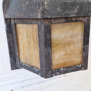 May include: A wall-mounted lantern featuring a dark, weathered metal frame with a distressed finish. The fixture includes square panels of translucent, amber-toned glass with a marbled texture, set within the sturdy, angular metal structure.