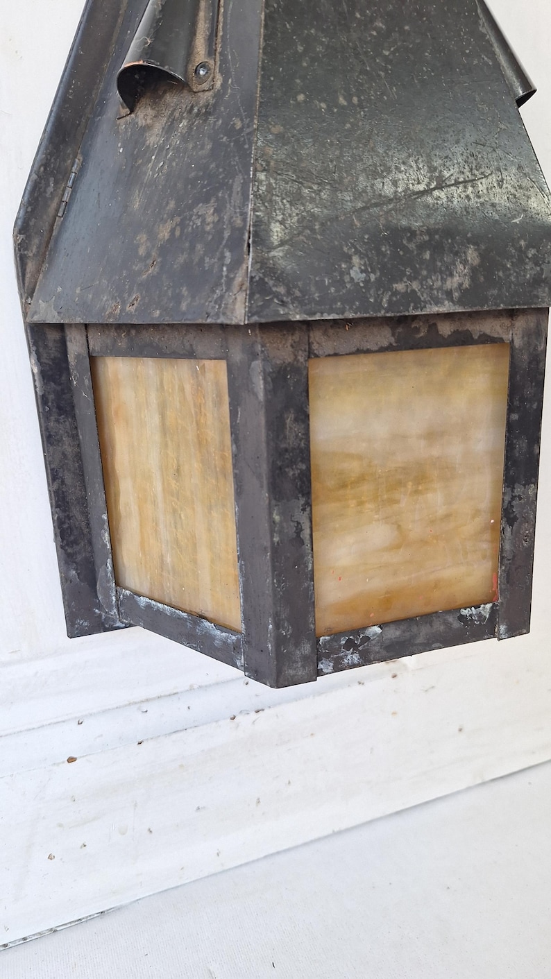 May include: A wall-mounted lantern featuring a dark, weathered metal frame with a distressed finish. The fixture includes square panels of translucent, amber-toned glass with a marbled texture, set within the sturdy, angular metal structure.