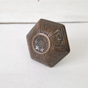 May include: A vintage, bronze-toned door knob with an ornate, floral design. The knob has a hexagonal shape with a circular center featuring a detailed, symmetrical pattern. The metal has a weathered appearance.