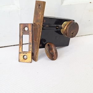 May include: An antique door lock set with a black metal body, brass-colored faceplates, and a keyhole. The set includes a rectangular strike plate, a long plate, and a small oval-shaped knob. The metal shows signs of age and wear.