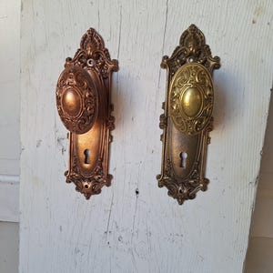 Puede incluir: Dos pomos de puerta antiguos con detalles ornamentados. Uno es de color bronce, el otro dorado. Ambos tienen pomos ovalados y cerraduras, montados sobre placas decorativas. Los pomos son probablemente de metal y vintage.