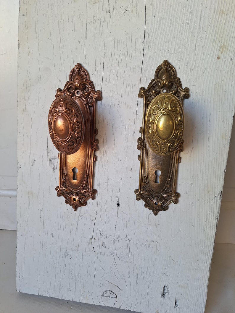 Puede incluir: Dos pomos de puerta antiguos con detalles ornamentados. Uno es de color bronce, el otro dorado. Ambos tienen pomos ovalados y cerraduras, montados sobre placas decorativas. Los pomos son probablemente de metal y vintage.