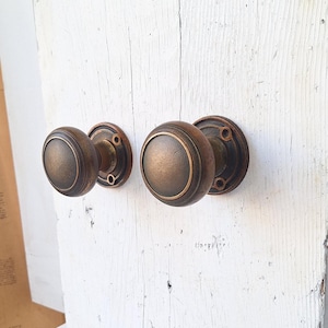 May include: Two antique bronze door knobs with a round design. The knobs have a dark bronze finish with a lighter bronze ring around the center. The knobs are mounted on a white wooden surface.