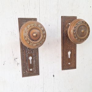 May include: Two antique bronze door knobs with matching rectangular backplates. The knobs are round with a detailed, textured pattern. The backplates feature an ornate design with a keyhole. The hardware is mounted on a white, weathered wooden surface.