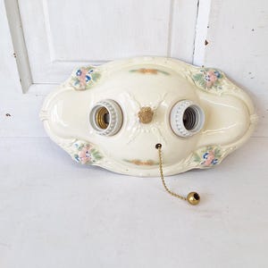 May include: Cream-colored, vintage-style ceiling light fixture with two bulb sockets. The fixture has floral accents in pink, blue, and orange. A gold-colored chain with a pull is attached.