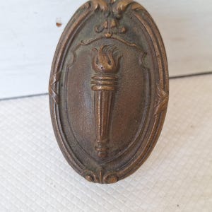 May include: Oval-shaped bronze-colored metal emblem featuring a torch within a shield design. The torch has a flame at the top and is surrounded by decorative flourishes. The emblem has an aged, antique appearance.