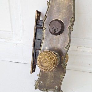 May include: Antique brass door hardware set featuring a decorative backplate with ornate detailing. The set includes a round doorknob with a floral design and a keyhole. The hardware is likely from the early 20th century.