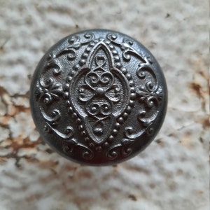 May include: A black metal knob with an ornate, raised design. The knob has a circular shape and a detailed, intricate pattern.