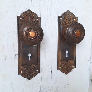 May include: Two antique bronze door knobs with ornate backplates. The knobs are round with a central copper-colored accent. The backplates feature decorative scrollwork and a keyhole. The hardware is mounted on a white, weathered wooden surface.