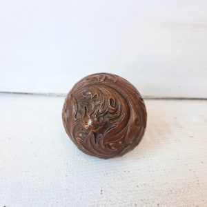 May include: A round, brown, wooden ornament with intricate carvings. The ornament has a floral design with a central motif that resembles a stylized flower or a bird.