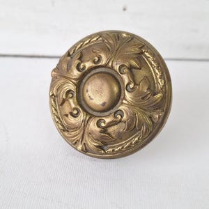 May include: Antique brass door knob with an ornate floral design. The round knob features a central circular element surrounded by detailed, swirling leaves and vines. The aged brass finish gives it a vintage aesthetic.