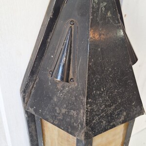 May include: A wall-mounted lantern with a weathered black metal frame and a pointed top. The fixture features translucent, amber-toned glass panels set within the metal structure. The metal surface shows signs of wear, scratches, and patina.