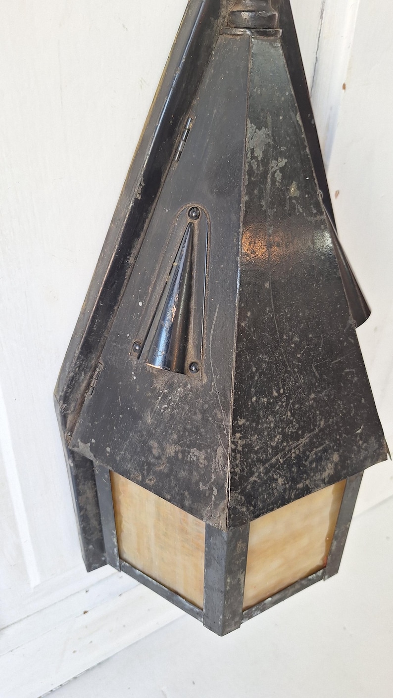 May include: A wall-mounted lantern with a weathered black metal frame and a pointed top. The fixture features translucent, amber-toned glass panels set within the metal structure. The metal surface shows signs of wear, scratches, and patina.