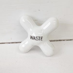 May include: A white, X-shaped ceramic faucet handle with the word "WASTE" printed in black. The handle has a glossy finish and is set against a white background.