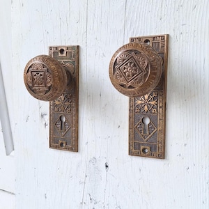 May include: Two antique brass door knobs with intricate designs and keyholes. The knobs are mounted on rectangular plates with ornate patterns.