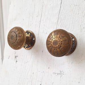 May include: Two antique brass door knobs with intricate floral designs. The knobs are round and have a raised center with a geometric pattern. The knobs are attached to round plates with a similar floral design.