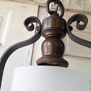 K&ouml;nnte beinhalten: Nahaufnahme einer Vintage-Pendelleuchte. Die Leuchte hat einen wei&szlig;en Lampenschirm und einen dunkelbronzenen Metallrahmen mit dekorativen Schn&ouml;rkeln. Das Licht h&auml;ngt an einer Kette, die an einer Deckenplatte befestigt ist.