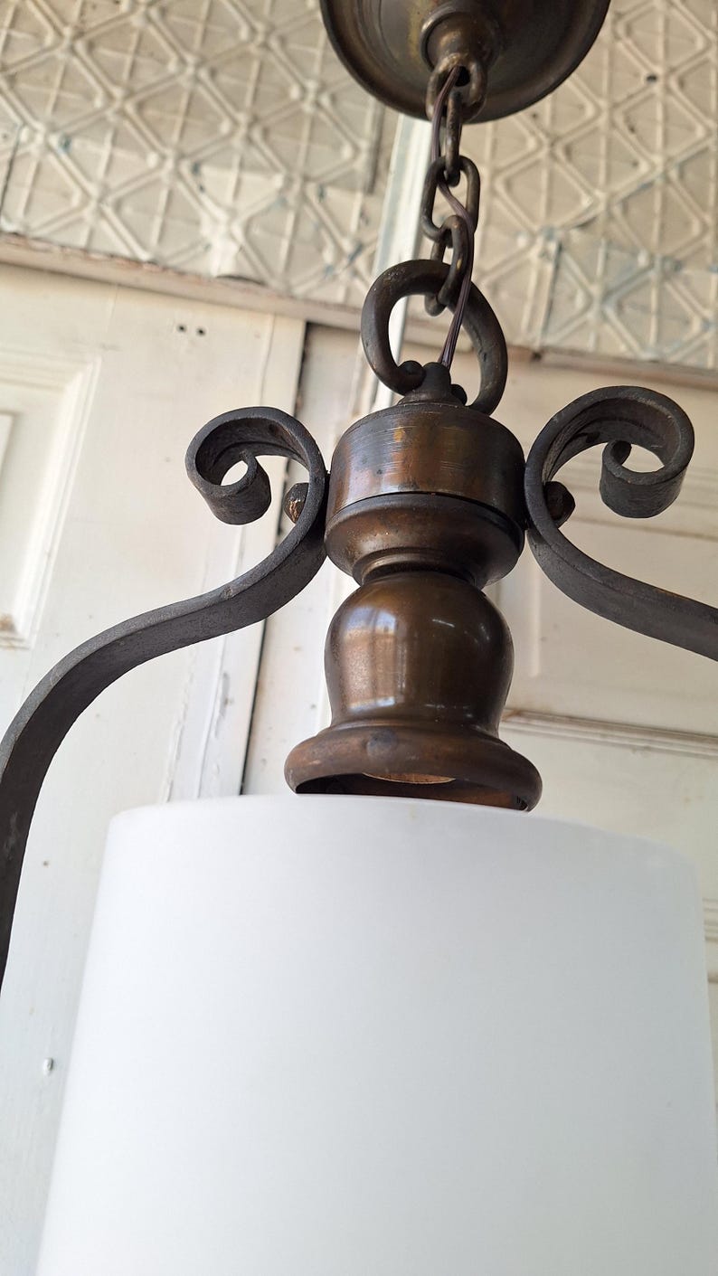 K&ouml;nnte beinhalten: Nahaufnahme einer Vintage-Pendelleuchte. Die Leuchte hat einen wei&szlig;en Lampenschirm und einen dunkelbronzenen Metallrahmen mit dekorativen Schn&ouml;rkeln. Das Licht h&auml;ngt an einer Kette, die an einer Deckenplatte befestigt ist.