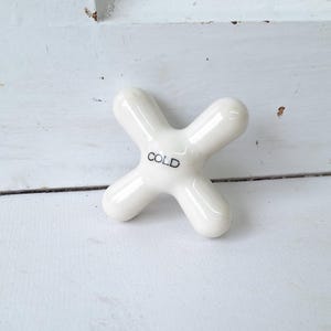 May include: A white, cross-shaped faucet handle with the word "COLD" printed in black. The handle is made of a glossy material and is set against a white, textured background. This vintage-style handle is a bathroom or kitchen fixture.