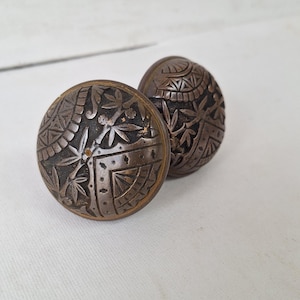 May include: Two antique bronze-colored cabinet knobs with detailed floral and geometric patterns. The knobs are round with a slightly domed shape, showcasing intricate designs. The background is a plain white surface.