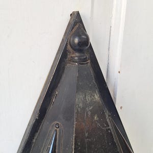 May include: A triangular black metal object with a pointed top and a spherical finial. The surface shows signs of wear and aging, featuring small conical protrusions on the sides and visible metal hinges along the edges of the frame.