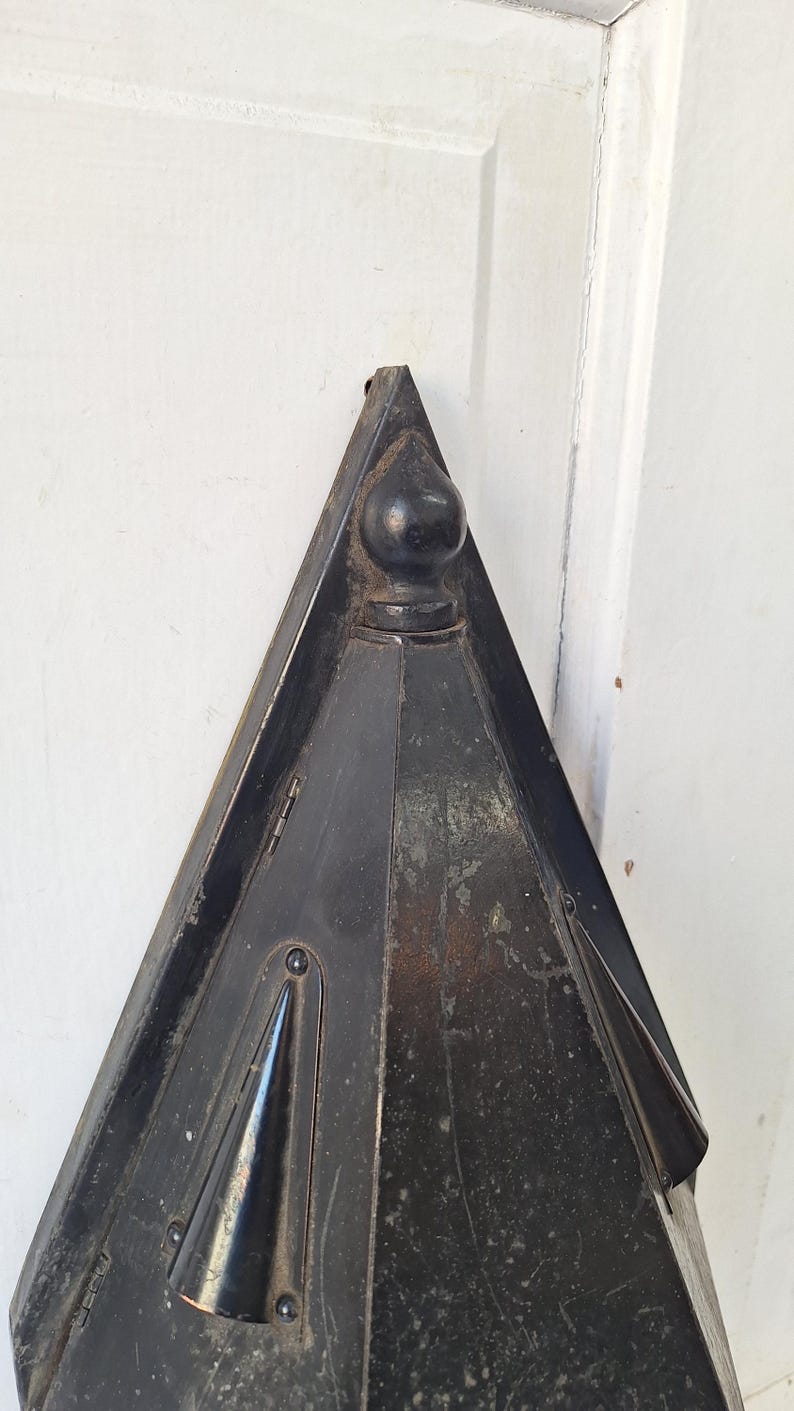 May include: A triangular black metal object with a pointed top and a spherical finial. The surface shows signs of wear and aging, featuring small conical protrusions on the sides and visible metal hinges along the edges of the frame.