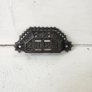 May include: Ornate, dark brown metal drawer pull with a decorative design. The pull has a semi-circular shape with a series of small circular cutouts along the top edge. The center features a detailed pattern with rectangular and floral elements.