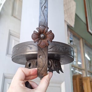 K&ouml;nnte beinhalten: Eine antike, zylindrische Leuchte mit einem Schirm aus Milchglas. Der Metallrahmen weist florale und Ranken-Details auf, mit einem markanten Blumenakzent. Die Leuchte wird von einer Hand gehalten, die ihr verziertes Design und ihre Vintage-&Auml;sthetik zeigt.