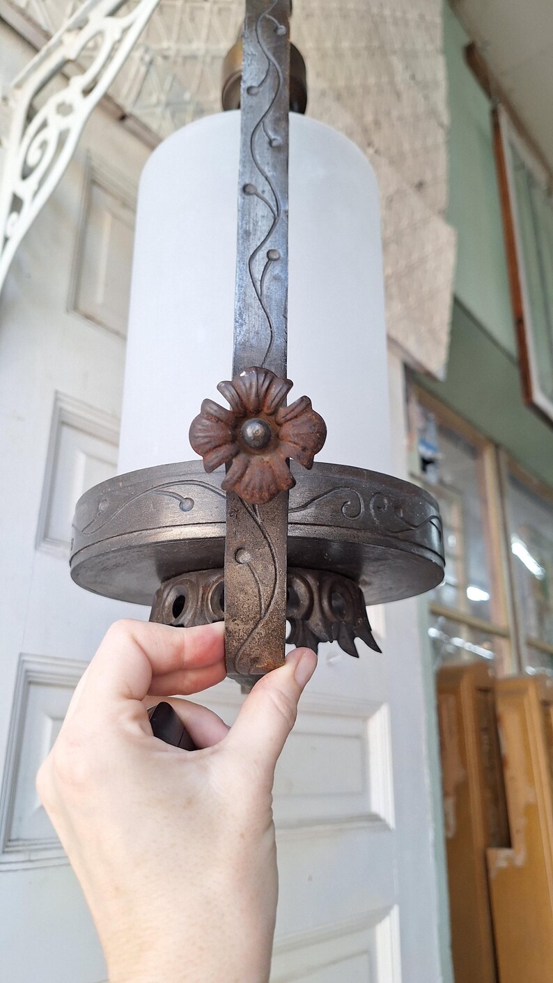 K&ouml;nnte beinhalten: Eine antike, zylindrische Leuchte mit einem Schirm aus Milchglas. Der Metallrahmen weist florale und Ranken-Details auf, mit einem markanten Blumenakzent. Die Leuchte wird von einer Hand gehalten, die ihr verziertes Design und ihre Vintage-&Auml;sthetik zeigt.