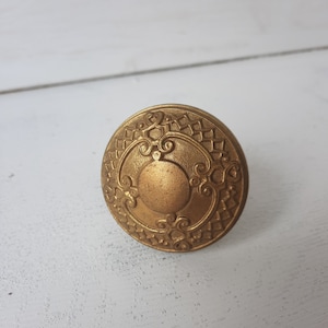 May include: A gold-toned metal knob with a decorative, ornate design. The knob has a circular shape with a raised center and intricate details around the edge.
