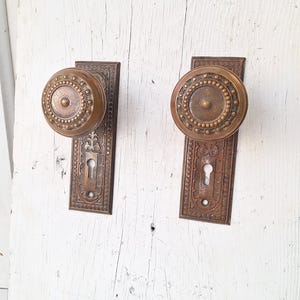 May include: Two antique bronze door knobs with matching backplates. The knobs are round with a beaded design around the perimeter. The backplates have an ornate, floral pattern and a keyhole. The hardware is mounted on a white, weathered wooden surface.