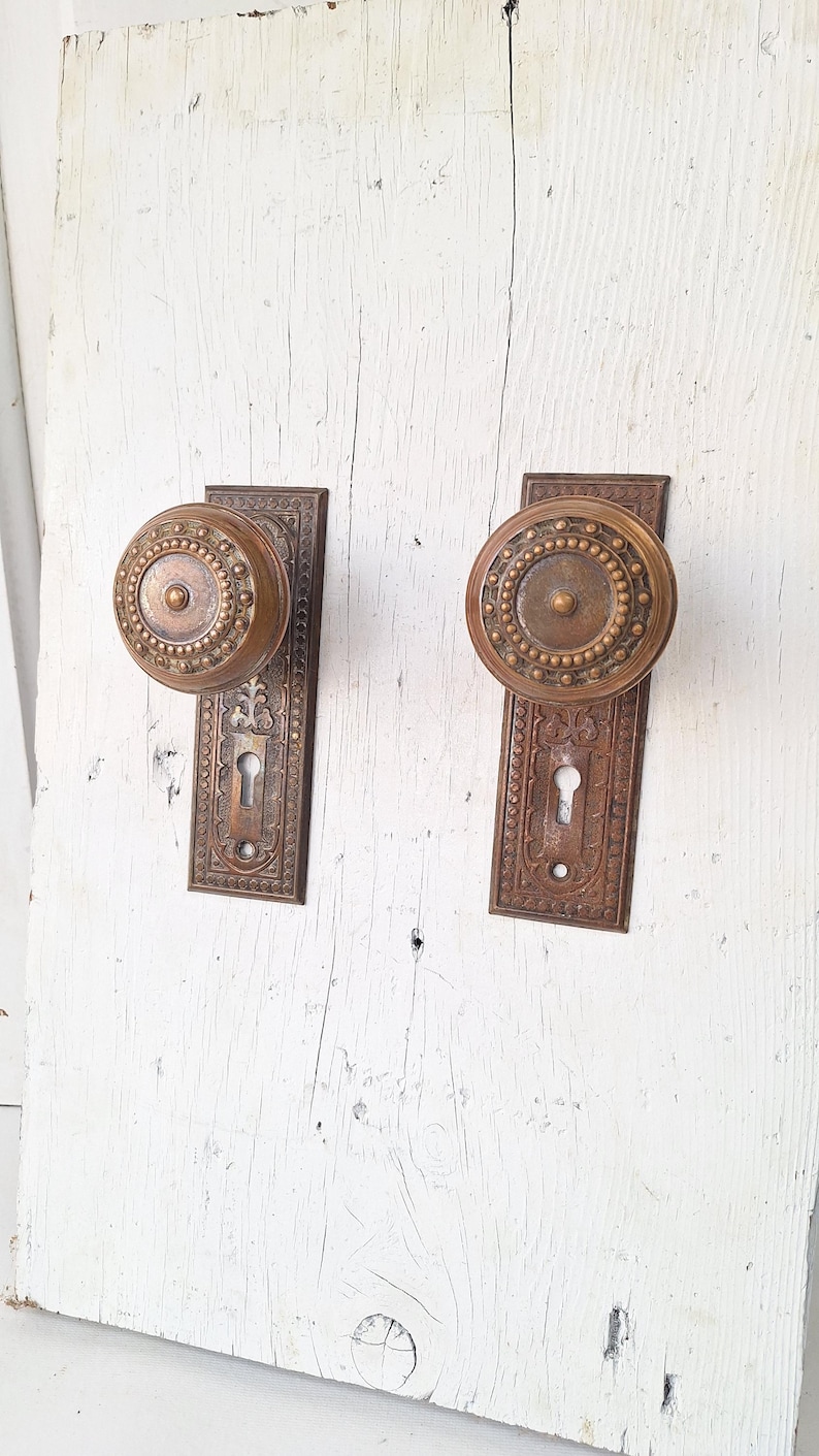May include: Two antique bronze door knobs with matching backplates. The knobs are round with a beaded design around the perimeter. The backplates have an ornate, floral pattern and a keyhole. The hardware is mounted on a white, weathered wooden surface.