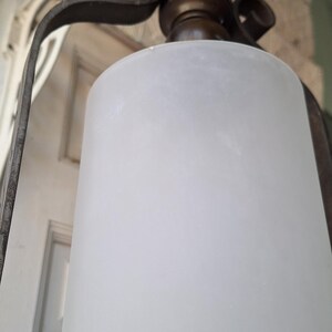 K&ouml;nnte beinhalten: Antike H&auml;ngelampe mit einem Schirm aus mattiertem Glas. Die Leuchte hat einen dunklen Metallrahmen mit dekorativen Schn&ouml;rkeln und eine messingfarbene Oberseite. Der zylindrische Schirm streut das Licht und erzeugt ein weiches Leuchten.