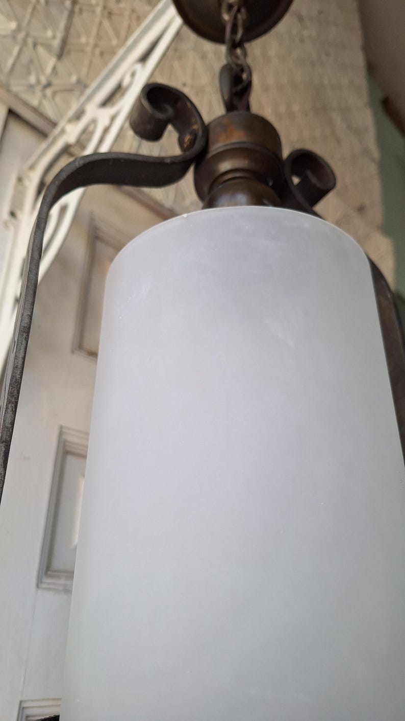K&ouml;nnte beinhalten: Antike H&auml;ngelampe mit einem Schirm aus mattiertem Glas. Die Leuchte hat einen dunklen Metallrahmen mit dekorativen Schn&ouml;rkeln und eine messingfarbene Oberseite. Der zylindrische Schirm streut das Licht und erzeugt ein weiches Leuchten.