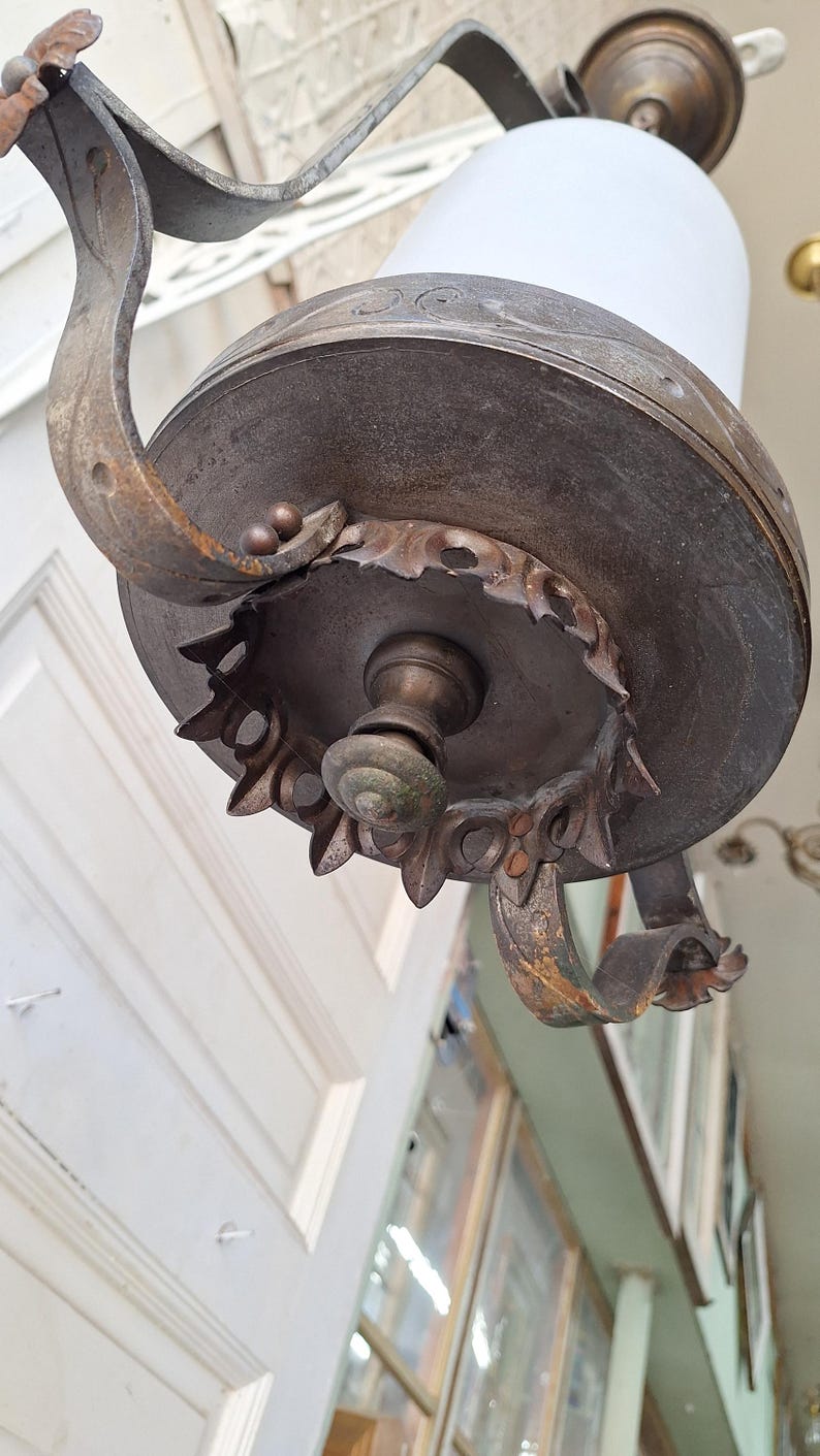 K&ouml;nnte beinhalten: Antike bronzefarbene H&auml;ngelampe mit wei&szlig;em Glasschirm. Die Leuchte verf&uuml;gt &uuml;ber verzierte, gebogene Metallarme und dekorative Details um die Basis. Das Design ist Vintage, mit einer klassischen, eleganten &Auml;sthetik.