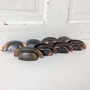 May include: A collection of seven semi-circular metal drawer pulls featuring a dark, weathered patina with visible copper-toned edges. Each pull has a curved handle design with two small circular mounting holes on the flat side extensions.