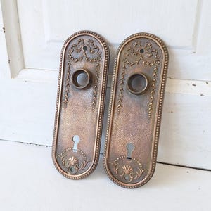 May include: Two antique bronze-colored door plates with ornate floral and scroll designs. Each plate has a keyhole and a circular opening. The plates are oval-shaped with a textured surface and a decorative border.