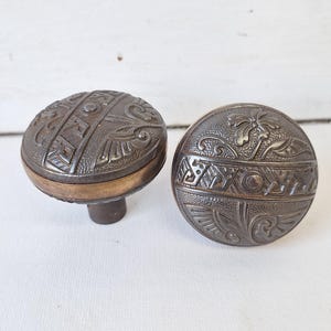 May include: Two antique door knobs with intricate designs. The knobs are made of dark metal with a brass trim. One knob features a floral design, while the other has a geometric pattern.