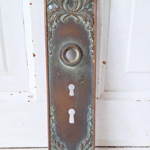 May include: Antique bronze door plate with ornate floral and scroll detailing. The rectangular plate features a keyhole and two key slots. The bronze has a patina with hints of green and brown, suggesting age and use.