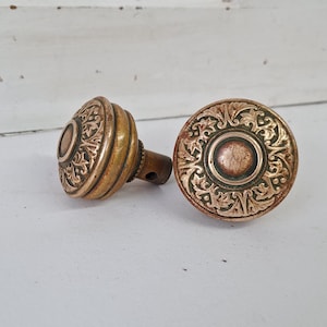 May include: Two antique brass door knobs with ornate floral designs. The knobs have a warm, aged patina, with detailed patterns on the round faces and cylindrical bases. One knob is positioned on its side, revealing the stem.
