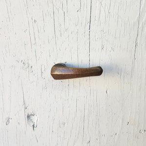 May include: A bronze-colored door handle with a textured surface is mounted on a white, weathered wooden door. The handle is horizontal and has a curved, ergonomic design, suggesting it is for pulling or opening a door.
