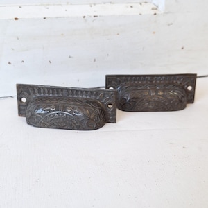 May include: Two antique bronze drawer pulls with ornate floral and geometric patterns. The pulls have a rectangular backplate with two holes for mounting and a curved, bowl-shaped handle. The hardware is likely for a cabinet or dresser.