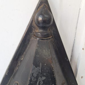 May include: A triangular black metal object with a pointed top and a central spherical finial. The surface shows signs of wear with visible paint chipping and texture. The structure features angled sides and a dark, weathered finish throughout.